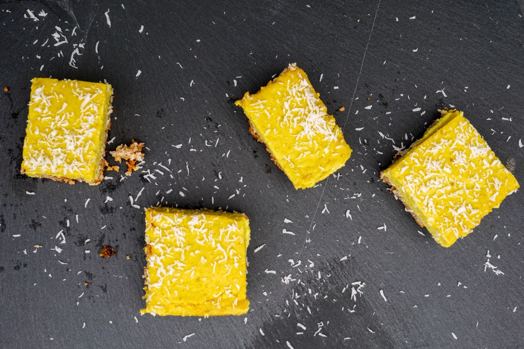 4 paleo turmeric lemon bars decorated with dessicated coconut on a black slate background