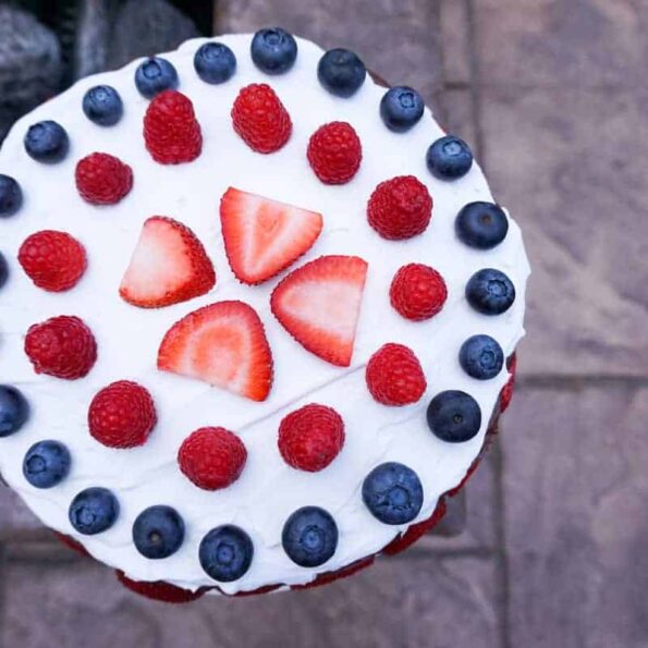 strawberries, raspberries and blue berries aranged in concentric circles on coconut whip cream on a chocolate cake near a fire pit.