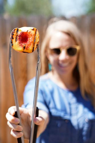 Girl in denim dress and gold glasses in front of wooden fence holding grilled peach in front of her with silver tongs. Cinnamon Grilled Peaches: a quick and healthy dessert perfect for summer Bar-B-Ques!