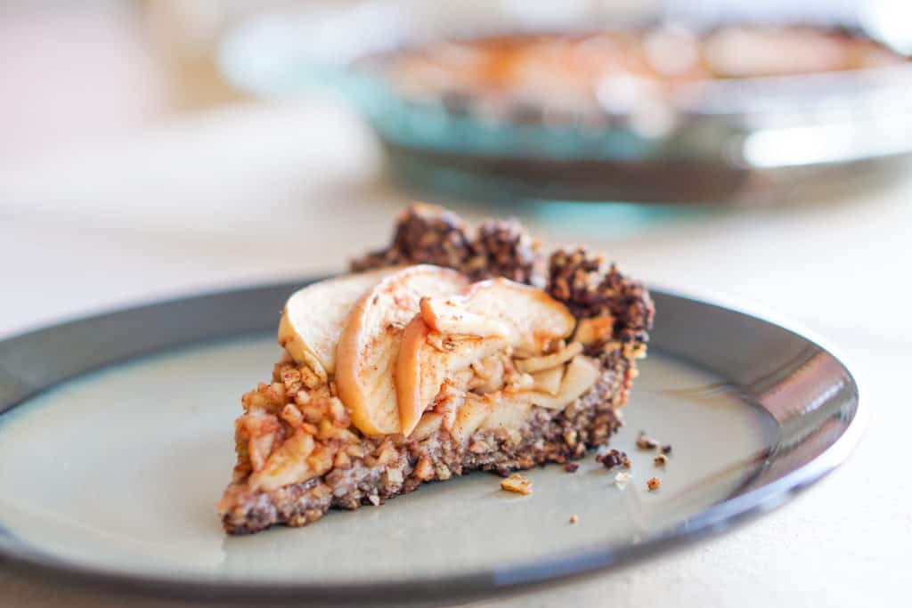 One slice of apple tart with chocolate drop cookie crust angled to the left on a green gray plate with a glass dish of gluten free dairy free apple tart int he background. 