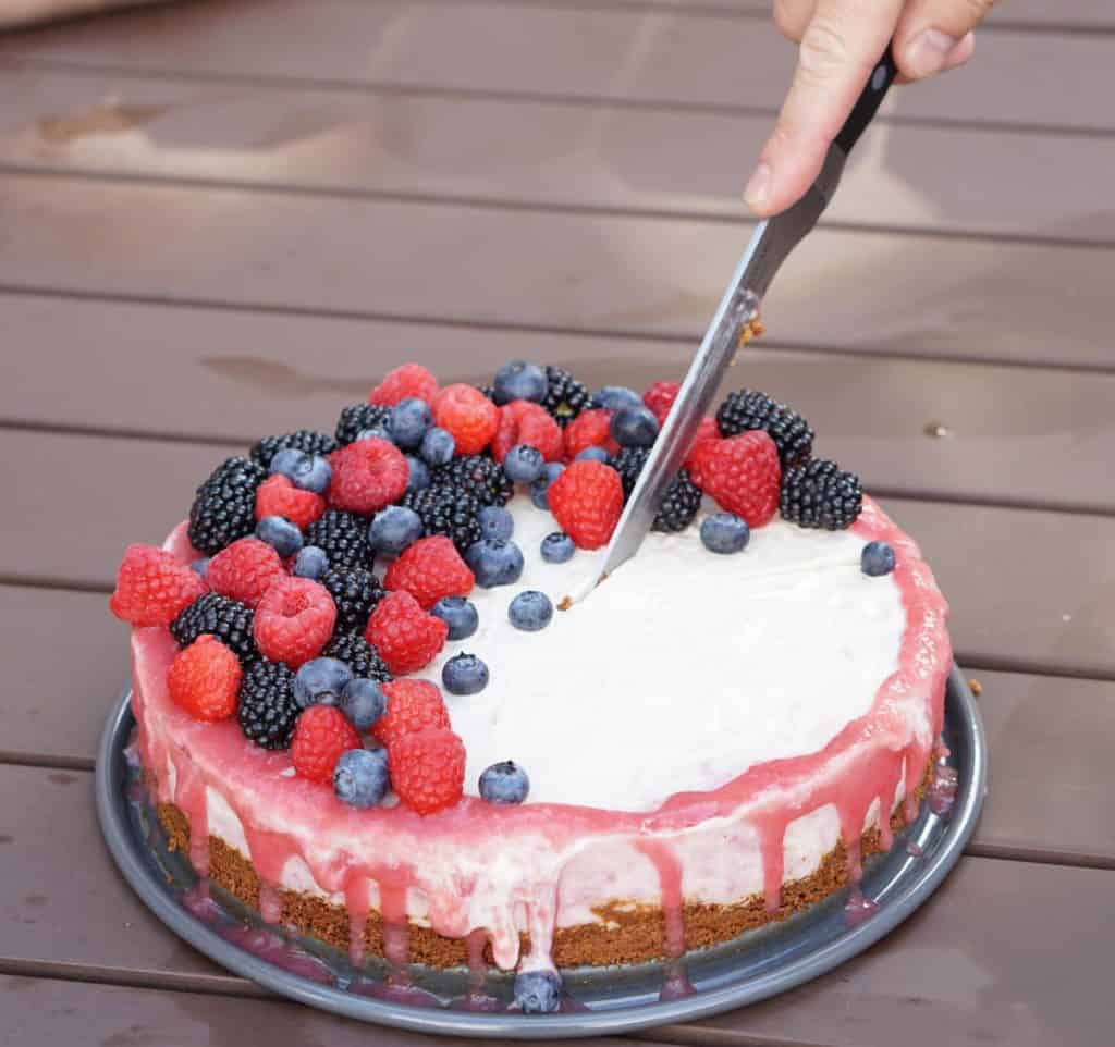 a person slicing into an ice cream cake with pink rhubarb drizzle and mixed berries and an almond cake crust on a picnic table at a BBQ