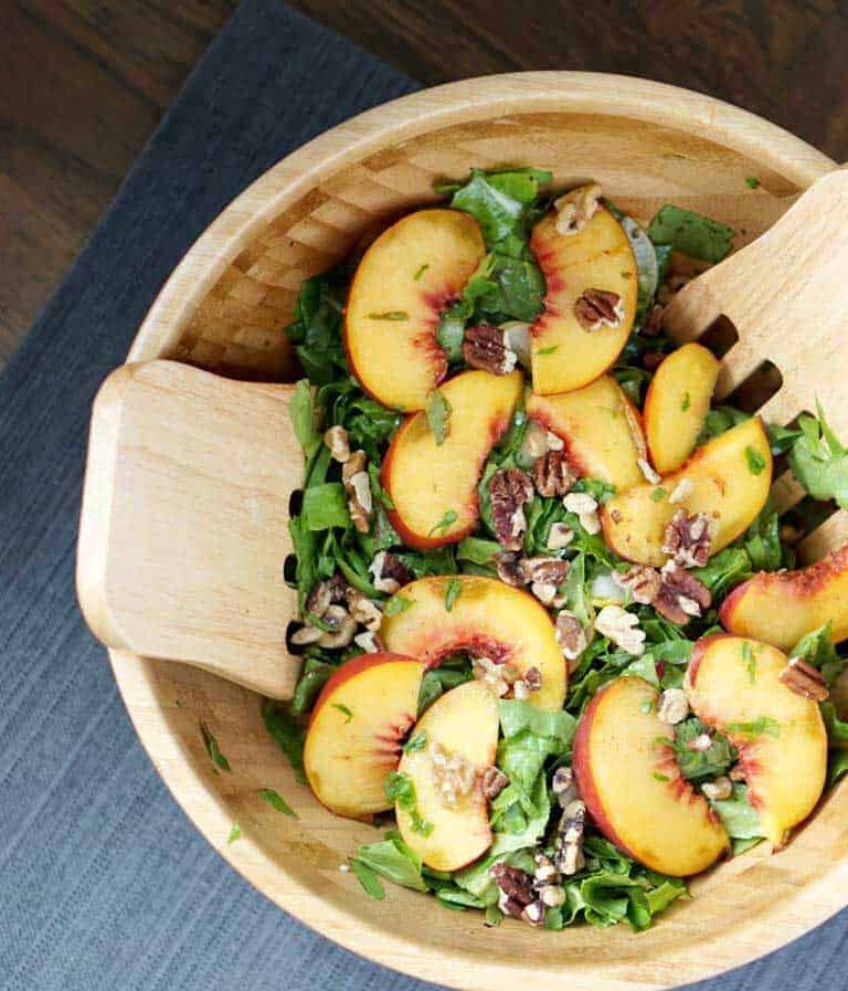 This toasted pecan, cucumber, basil, and peach salad in a large wooden bowl with wooden salad spoons on a darker wooden table is super fresh and incorporates summer and fall flavors into one great gluten and dairy free dish. It's also super quick and sugar-free