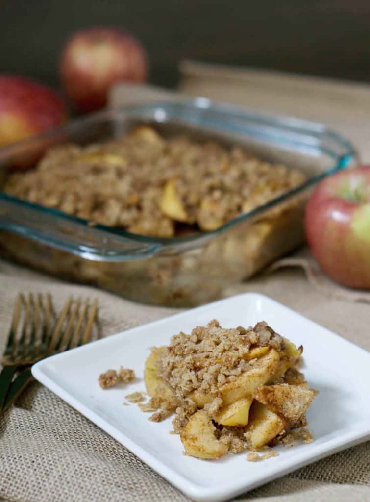 Cinnamon and clove covered apples melting into a paleo, gluten free crumble crust on a white plate next to two bronze forks with fresh apples and a glass tray of apple crisp in the background on burlap canvas tablecloth