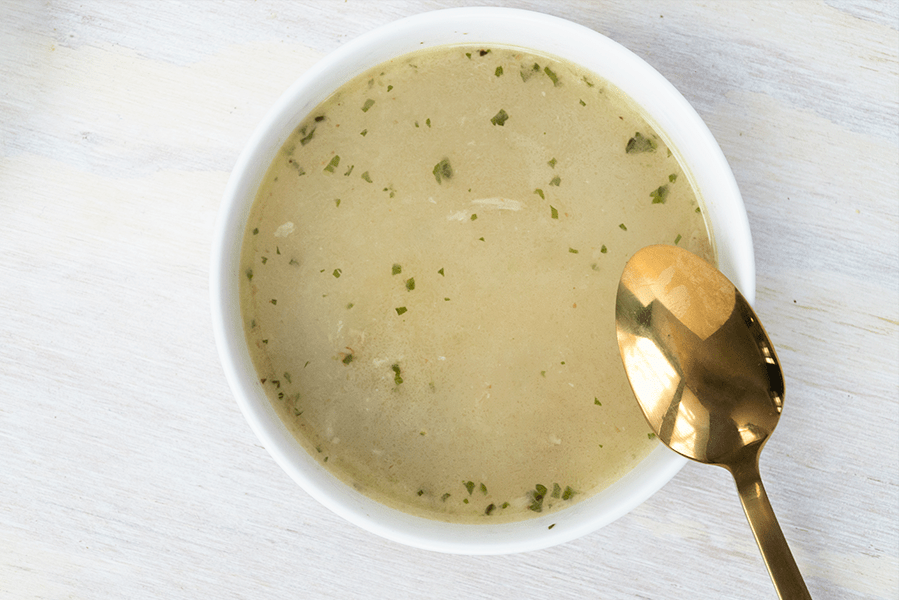 One bowl of chrsitmas goose stew. A yellow goose broth thickened with vegetables and finished with small pieces of fresh green parsley on a white table in a white bowl with a golden spoon about to dive in