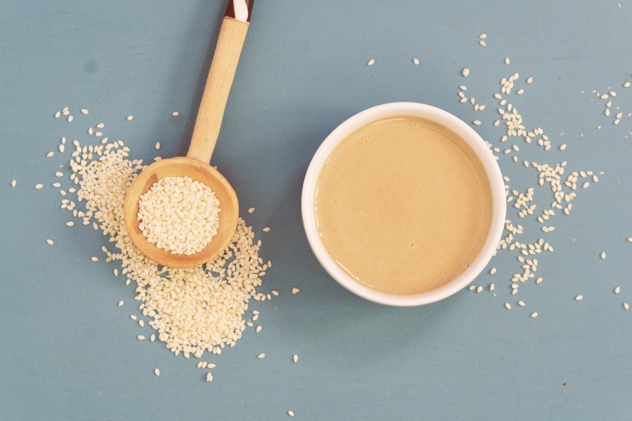 White bowl of tahini with wooden spoon overflowing with sesame seeds on vintage blue showing what is tahini, how it's made, tahini benefits, and how to use it.