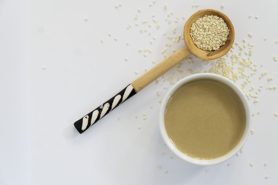 White bowl of tahini with wooden spoon overflowing with sesame seeds showing what is tahini, how it's made, tahini benefits, and how to use it.