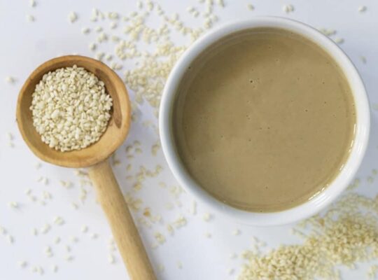 White bowl of tahini with wooden spoon overflowing with sesame seeds showing what is tahini, how it's made, tahini benefits, and how to use it.
