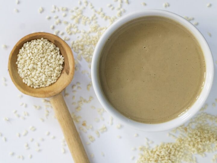 White bowl of tahini with wooden spoon overflowing with sesame seeds showing what is tahini, how it's made, tahini benefits, and how to use it.