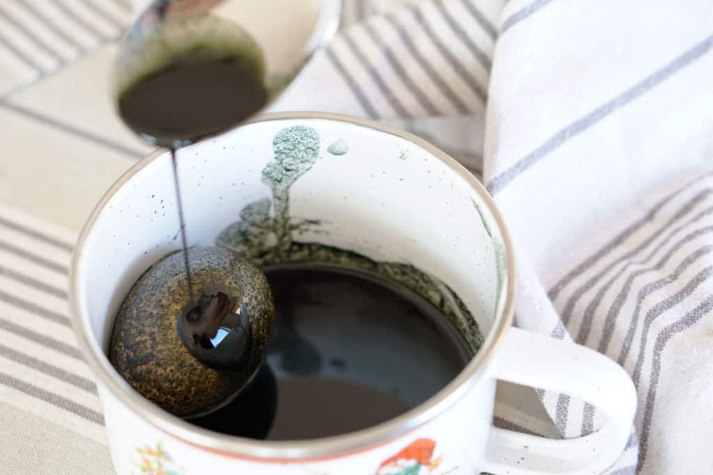 Green food coloring dripping off of a spoon onto an Easter Egg in green dye in a white mug.