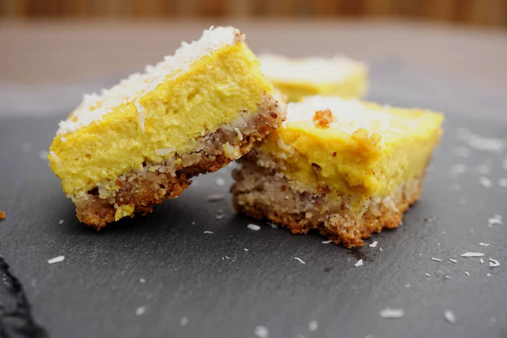 Yellow lemon bars with nutty crust stacked on top of eachother on a black slate tray with a wooden background