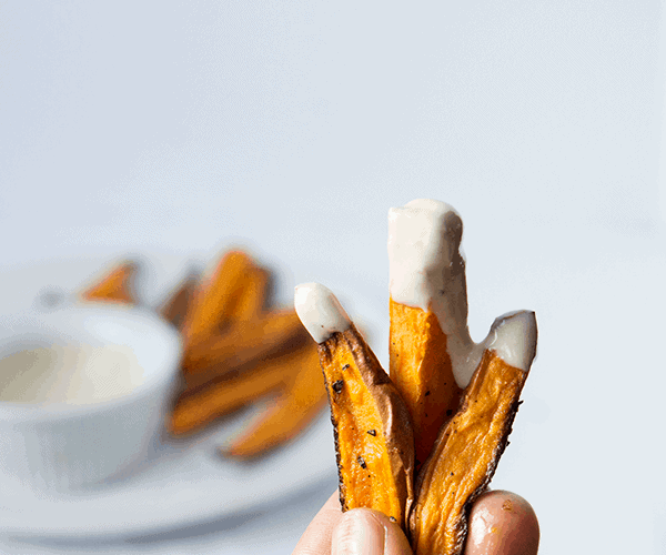 hand holding 3 sweet potato fries dipped in whipped tahini sauce with sauce and fries in the background.
