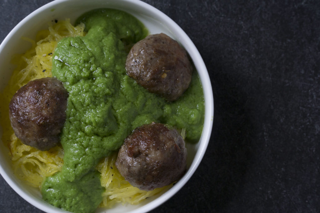 Green sauce smothered meatballs on spaghetti squash in white bowl on dark marble and poured concrete surface