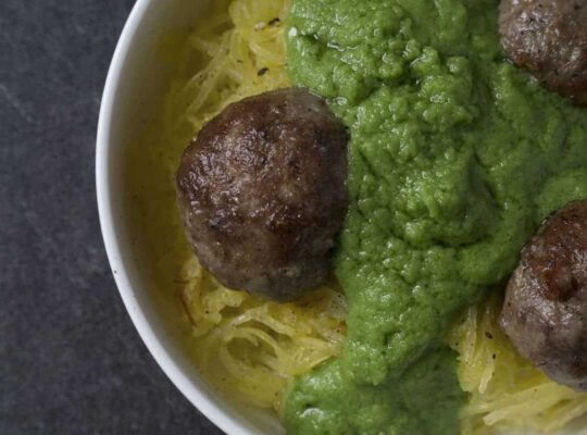 Green sauce smothered meatballs on spaghetti squash in white bowl on dark marble and poured concrete surface