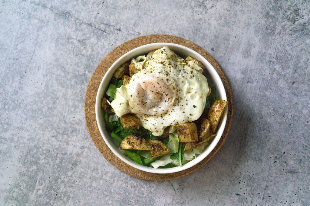 White bowl on cork circle on cement with sauteed cabbage and turnip greens with harissa roasted turnips and broth poached eggs