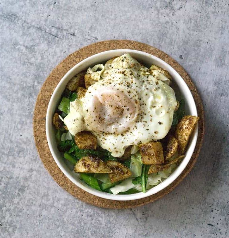 White bowl on cork circle on cement with sauteed cabbage and turnip greens with harissa roasted turnips and broth poached eggs