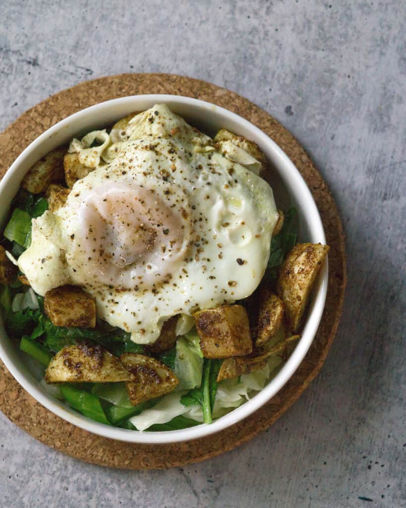 White bowl on cork circle on cement with sauteed cabbage and turnip greens with harissa roasted turnips and broth poached eggs