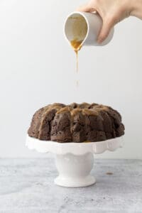 hand pouring coconut cardamom glaze onto chocolate chai bundt cake on white cake stand on cement table with white backdrop