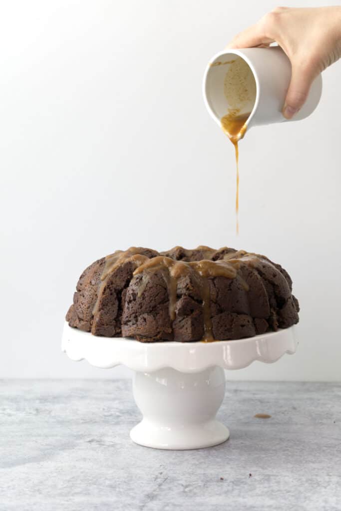 hand pouring coconut cardamom glaze onto chocolate chai bundt cake on white cake stand on cement table with white backdrop