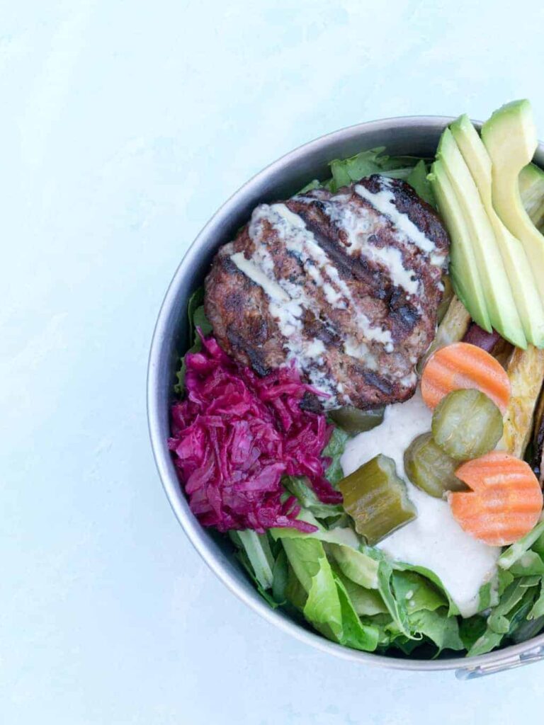 Paleo Elk Burger Bowl in Tin bowl on cement background: mixed greens topped with cashew cream, pickled cucumbers and carrots, roasted sweet potato fries, red sauerkraut, elk burger and tahini