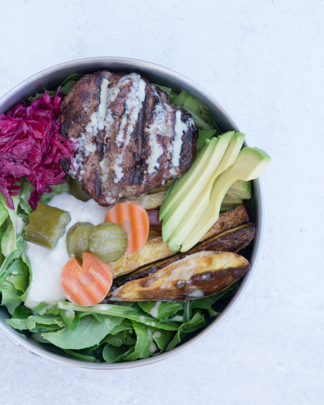 Paleo Elk Burger Bowl in Tin bowl on cement background: mixed greens topped with cashew cream, pickled cucumbers and carrots, roasted sweet potato fries, red sauerkraut, elk burger and tahini