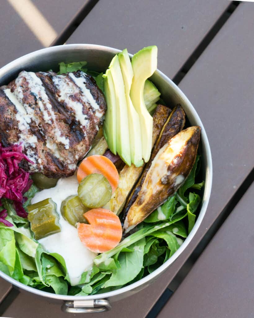 Paleo Elk Burger Bowl in Tin bowl on wooden picnic table: mixed greens topped with cashew cream, pickled cucumbers and carrots, roasted sweet potato fries, red sauerkraut, elk burger and tahini