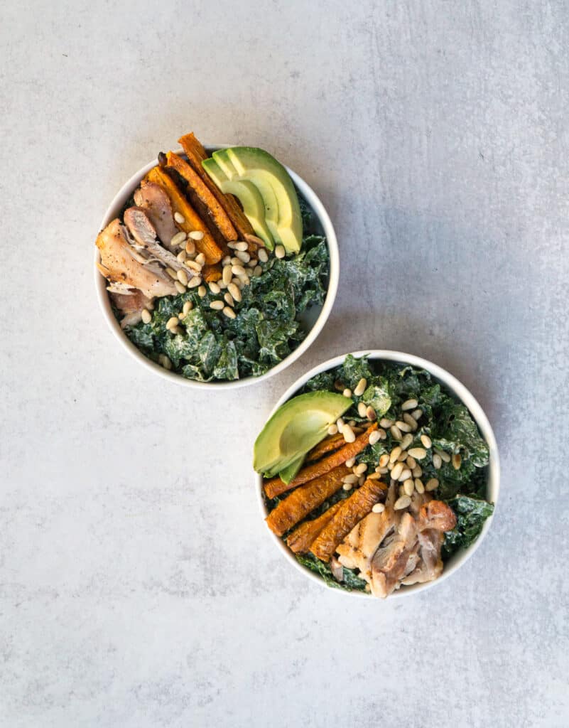 Superfood Kale Caesar salad in 2 white bowls with avocado, carrot fries, pine nuts and chicken thighs with vegan Caesar dressing