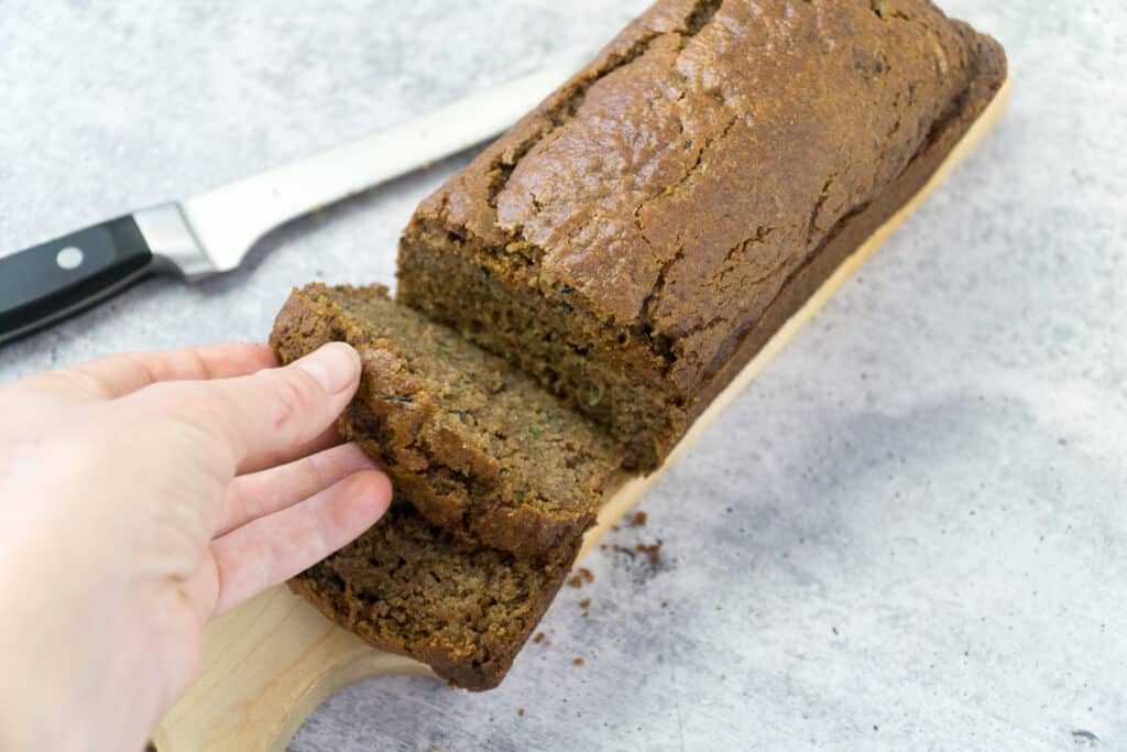 Grabbing a slice of gluten-free spiced zucchini bread from a cutting board, sitting by a knife