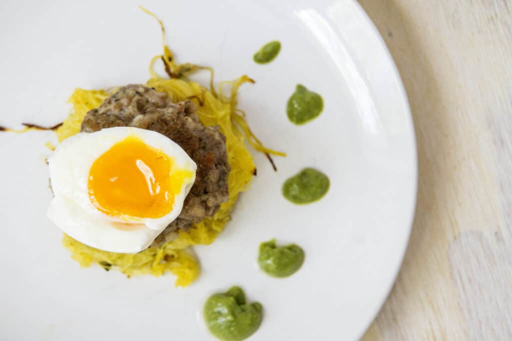 1/2 soft boiled egg over a sausage pattie and spaghetti squash nest with dotted green sauce on white plates