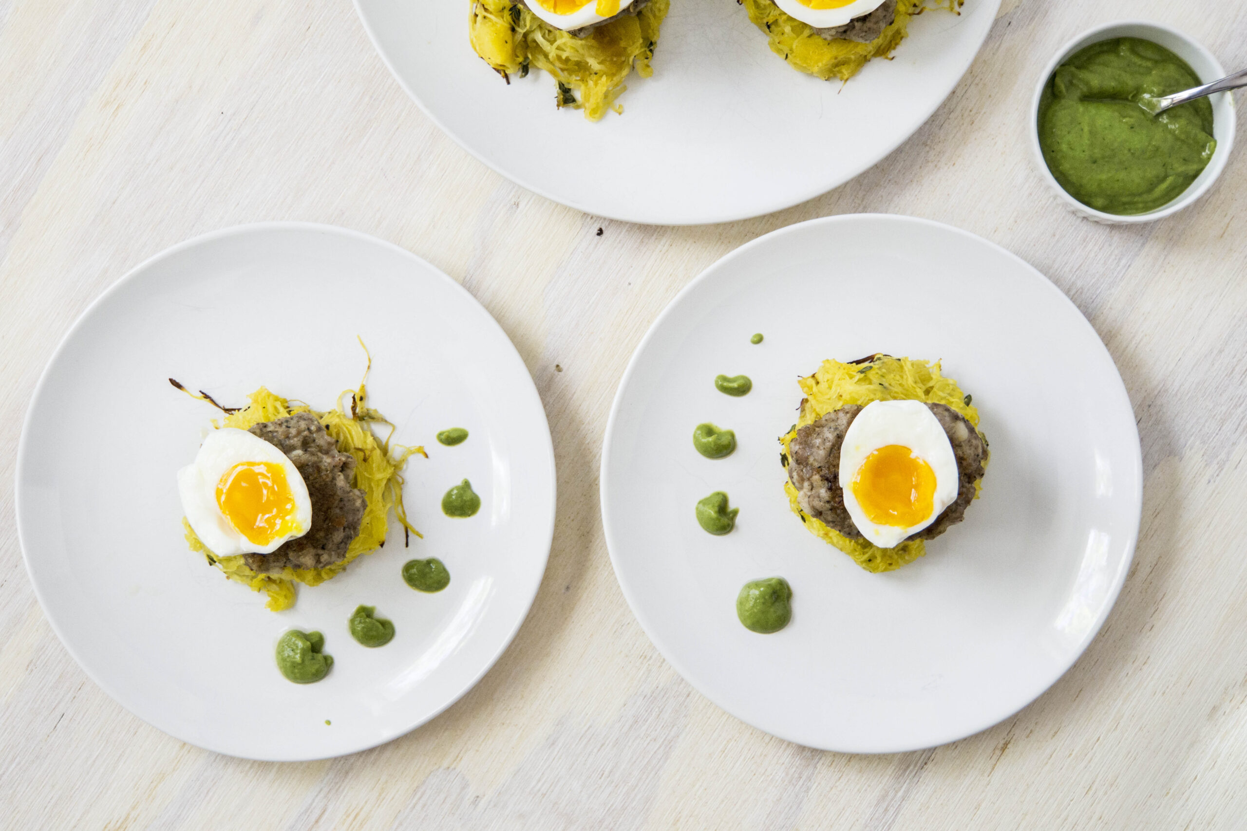1/2 soft boiled egg over sausage patties and spaghetti squash nests with dotted green sauce on white plates