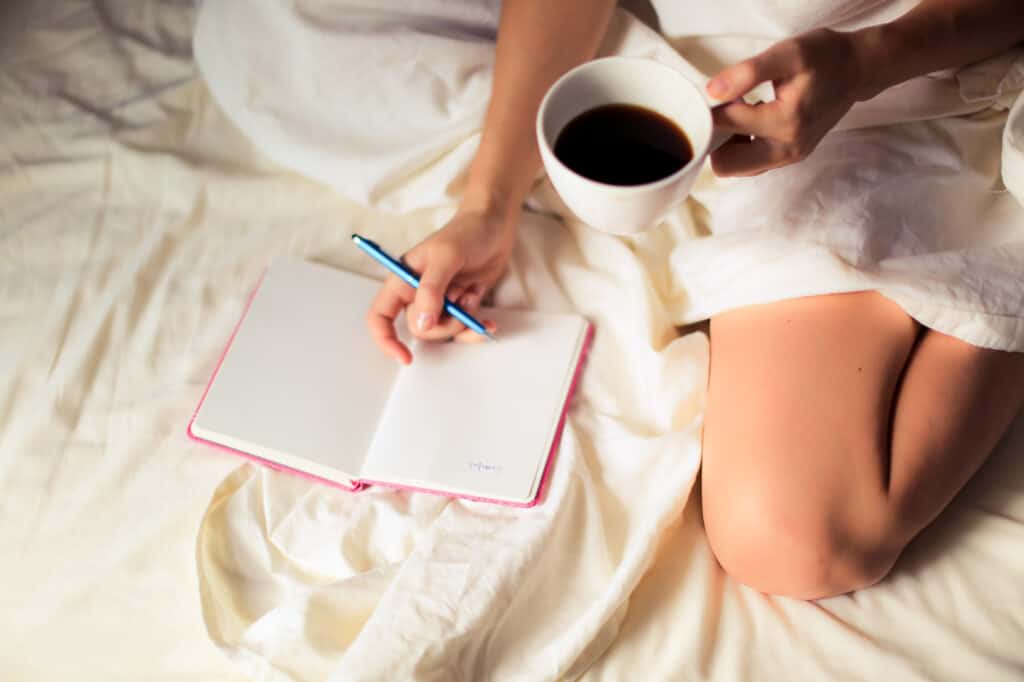 Woman in bed journaling in a pink notebook with a cup of coffee