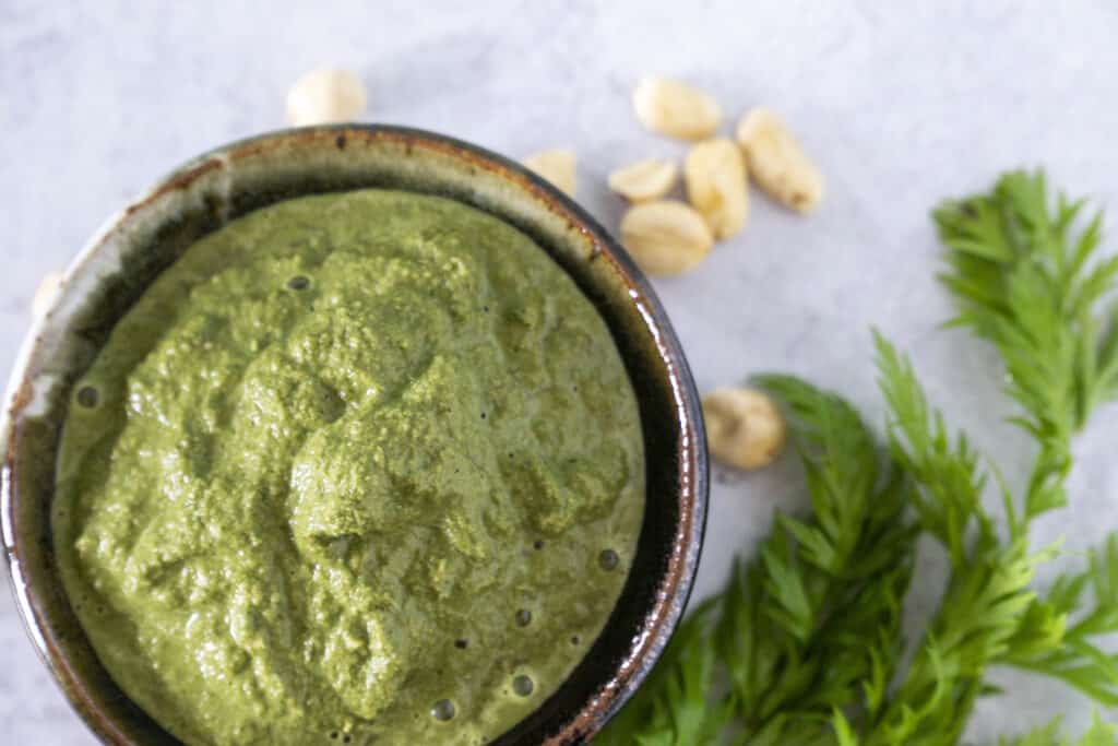 Green Carrot Top Pesto in ceramic bowl on counter with carrot tops and peanuts on the counter