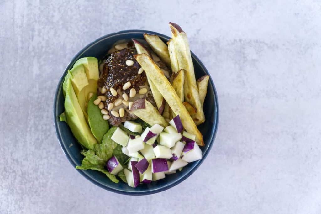 Overhead view of A hand grabs a Fig Tahini Burger bowl with sweet potato fries, kohlrabi and avocado