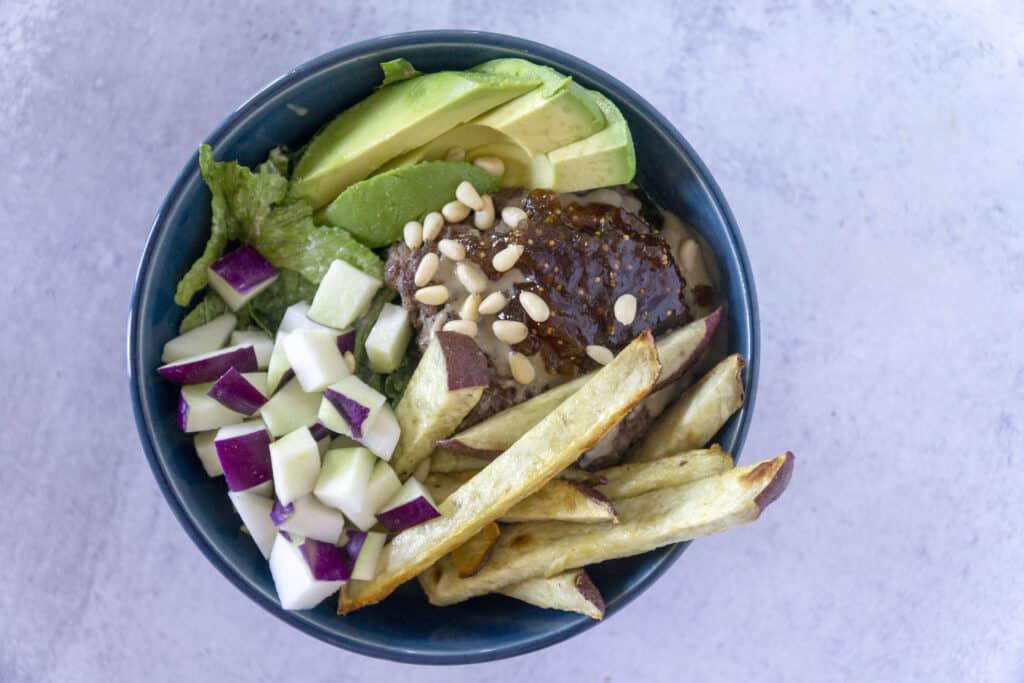 Overhead view of A hand grabs a Fig Tahini Burger bowl with sweet potato fries, kohlrabi and avocado 