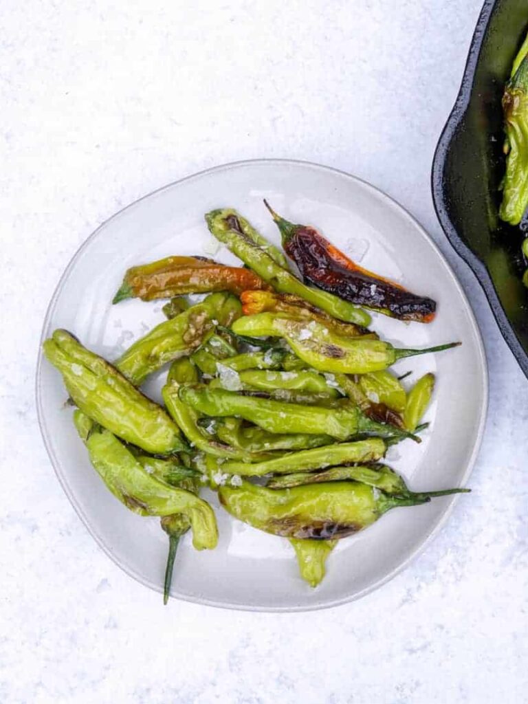 Blistered Shishito peppers on a plate next to a cast iron of peppers