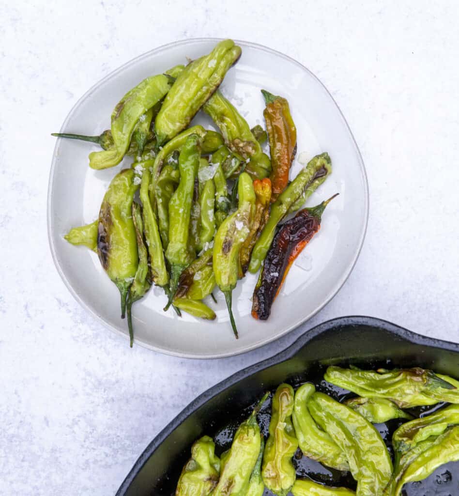 Blistered Shishito peppers on a plate next to a cast iron of peppers tall