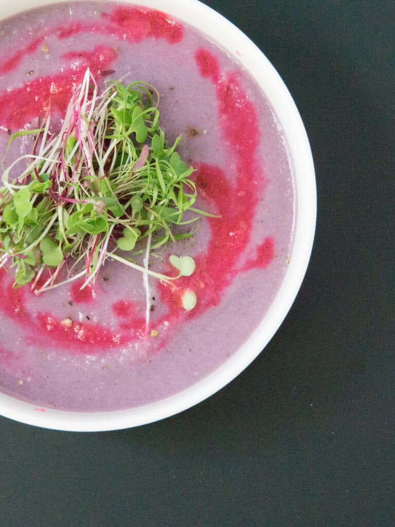 white bowl of bright purple soup with fuchsia drizzle and micro greens on black table