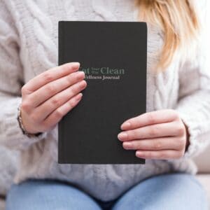 Woman holding eat your way clean wellness journal, sitting down above her lap. In Denim and cozy sweater