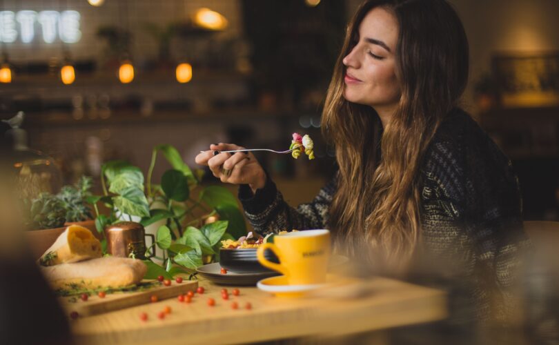Contented woman smiling and eating in peace - at a wooden dinner table with lots of veggies, a big plate of food and a bright yellow cup - overall sunny and joyful vibe
