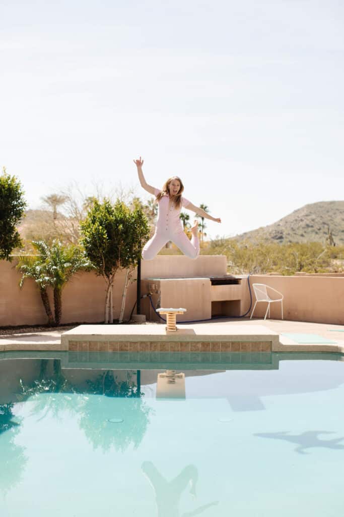 Woman in pink carpenter romper jumping into pool form diving board in desert