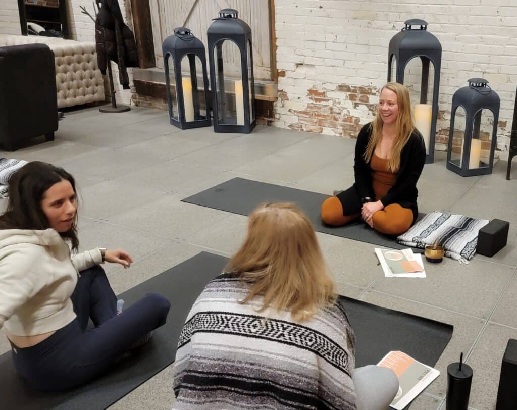 blond woman in orange and black yoga outfit teaching mindfulness to ladies on yoga mats in blankets with large candles in background