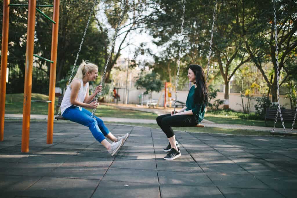 2 women one blondi n athletic gear, one brunette in casual wear, facing each other on swing set in otherwise empty park