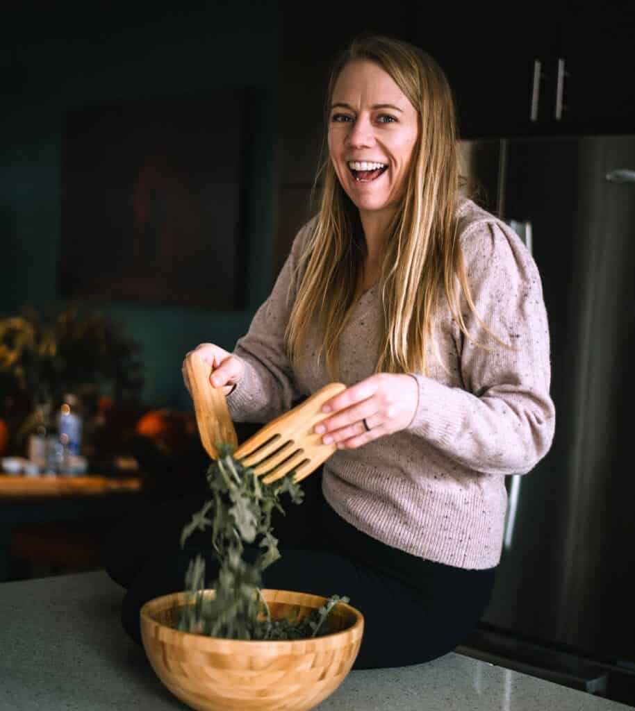 Blonde women with long hair and pink sweater sitting on counter mixing arugula salad with wooden salad forks and wooden salad owl