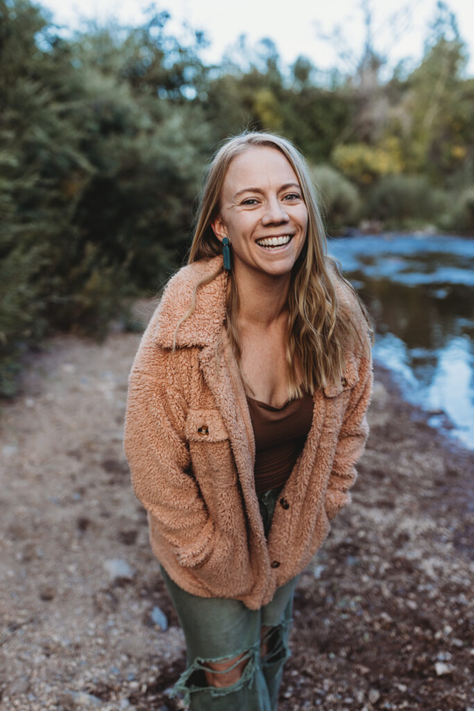Blond women in colorado forest near river with tan tank, pink furry coat and green jeans, hands together leaning towards you