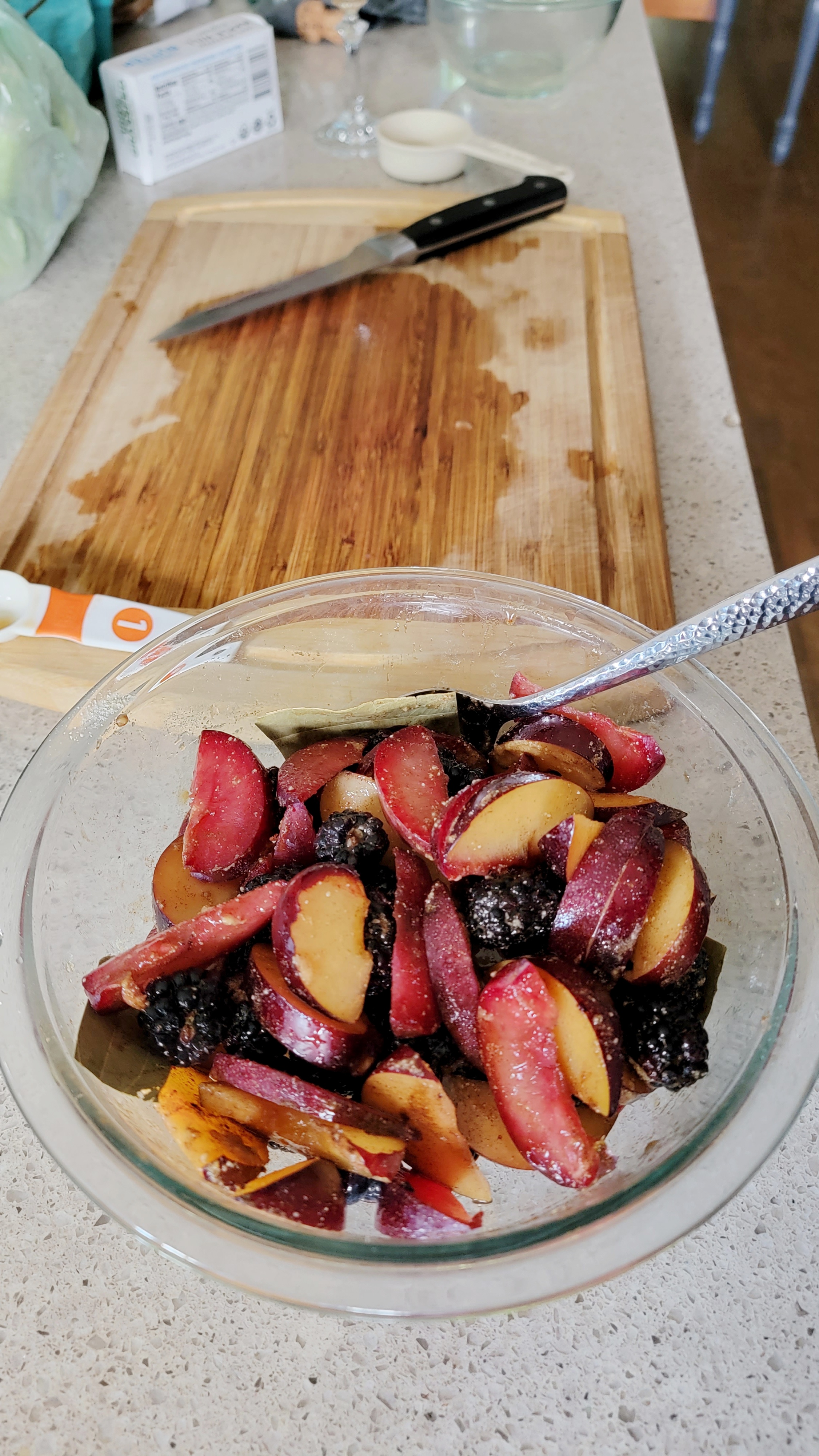 A glass bowl sits on a counter with wedged plums, blackberries, sugar and bay leaves mixed together. It sits near a cutting board, knife and other kitchen baking equipment