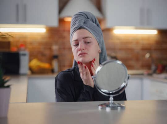 woman feeling shock while looking problem with skin condition after illness by mini mirror. Conceptual shot of Acne and Problem Skin on female face. with a towel covering wet hair
