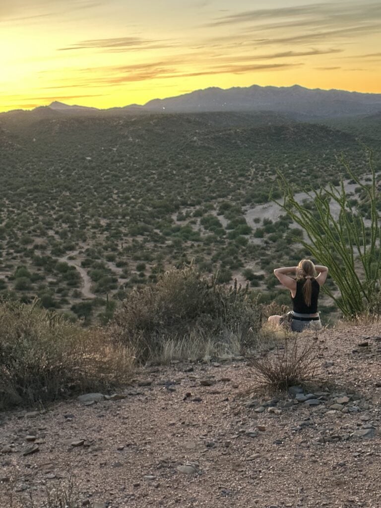 Girl in black tank and green sweat shorts, sitting cross legged in the desert with hands on her head, sun is setting behind bluffs