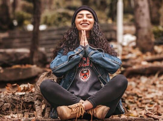 Women in sweats and jean jacket with curly hair loose under a beanie, sitting cross leged in the forest on the ground with hands in prayer pose, eyes closed, meditating