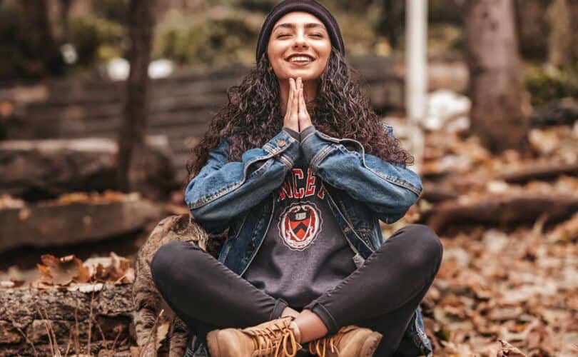 Women in sweats and jean jacket with curly hair loose under a beanie, sitting cross leged in the forest on the ground with hands in prayer pose, eyes closed, meditating