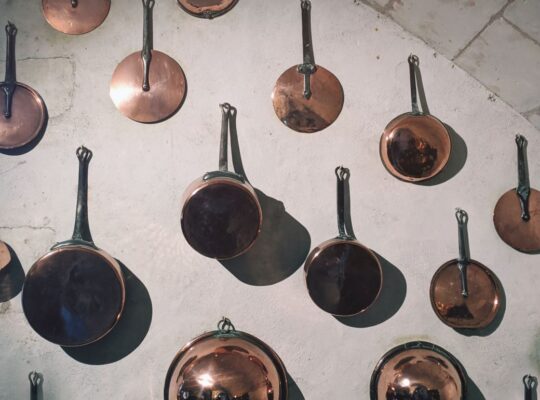 varios pots, mostly copper, some steel, hanging on a white wall by nails - spaced about 8 inches apart