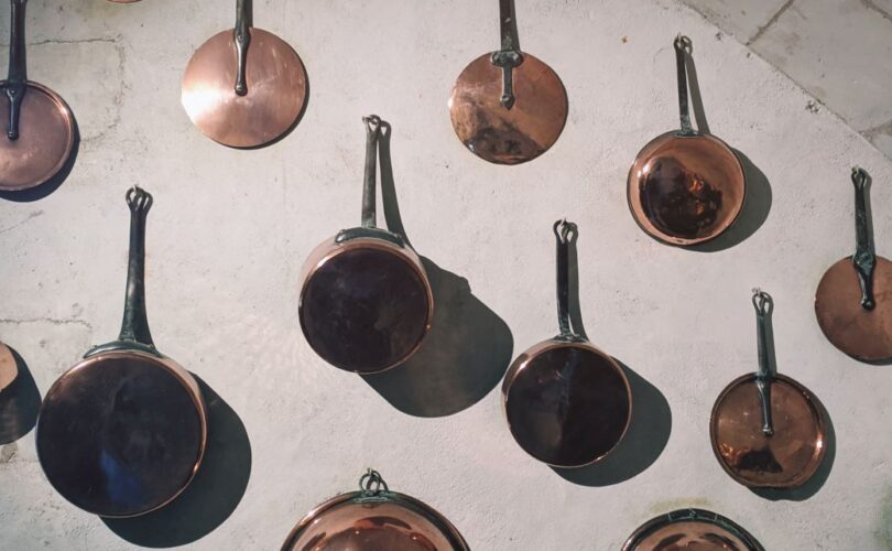 varios pots, mostly copper, some steel, hanging on a white wall by nails - spaced about 8 inches apart